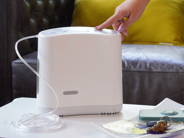 Side View Of The O2 Concepts Oxilife Liberty Portable Oxygen Concentrator In A Home Setting Shown With A Hand Pushing A Button On The Device