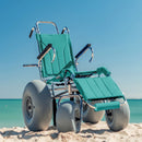Wheeleez Sandcruiser All-Terrain Beach Wheelchair Front View In Aruba Teal Color With Large Balloon Wheels, Positioned On A Sandy Beach With Ocean In The Background. 