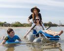 Wheeleez Sandcruiser All-Terrain Beach Wheelchair Front View In Mediterranean Blue Color With Large Balloon Wheels, Positioned In The Water With One Woman Sitting In The Sandcruiser, One Woman Pushing The Wheelchair And A Child Swimming With A Tent In The Background On A Sunny Day.