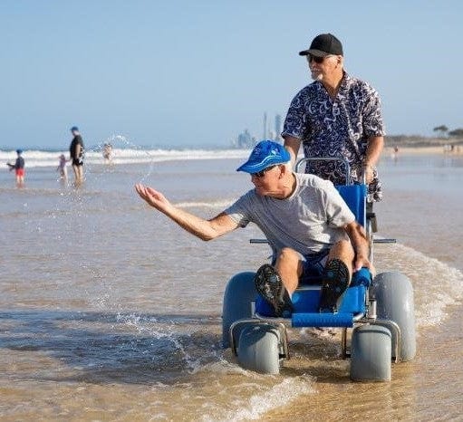 Wheeleez Sandcruiser All-Terrain Beach Wheelchair Front View In Mediterranean Blue Color With Large Balloon Wheels, Positioned In The Water Along The Beach Front With One Man Sitting In The Sandcruiser And One Man Pushing The Wheelchair On A Sunny Day.