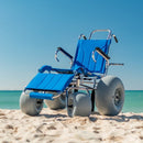 Wheeleez Sandcruiser All-Terrain Beach Wheelchair Front View In Mediterranean Blue Color With Large Balloon Wheels, Positioned On A Sandy Beach With Ocean In The Background. 