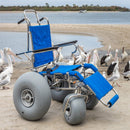 Wheeleez Sandcruiser All-Terrain Beach Wheelchair Front View In Mediterranean Blue Color With Large Balloon Wheels, Positioned On A Sandy Beach With Seagulls And Ocean In The Background. 