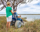 Wheeleez Sandcruiser All-Terrain Beach Wheelchair Rear View In Mediterranean Blue Color With Large Balloon Wheels And RoboCup Holder, Positioned On A Sandy Beach With Man Sitting In The Wheelchair While A Woman Stands Behind The Wheelchair Holding The Handles With Ocean