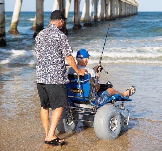 Wheeleez Sandcruiser All-Terrain Beach Wheelchair Rear View In Mediterranean Blue Color With Large Balloon Wheels, Positioned On A Sandy Beach With Man Sitting In The Wheelchair Fishing While A Man Stands Behind The Wheelchair Holding The Handles With Ocean In The Background