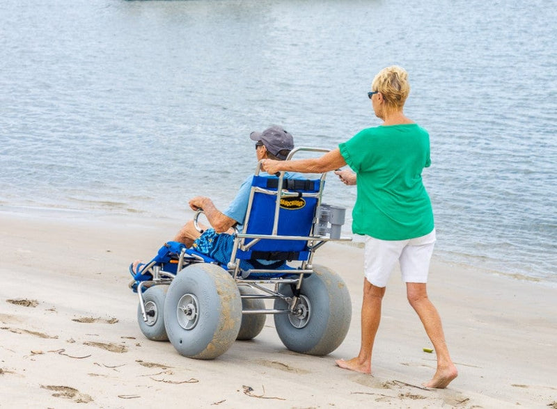 Wheeleez Sandcruiser All-Terrain Beach Wheelchair Rear View In Mediterranean Blue Color With Large Balloon Wheels And RoboCup Holder, Positioned On A Sandy Beach With Man Sitting In The Wheelchair While A Woman Pushes The Wheelchair And Ocean In The Background.  