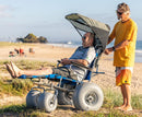 Wheeleez Sandcruiser All-Terrain Beach Wheelchair Side View In Mediterranean Blue Color With Large Balloon Wheels And Canopy, Positioned On A Sandy Beach With One Man Sitting While Another Man Standing Behind The Wheelchair Holding The Handles And Ocean In The Background.  