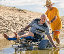 Wheeleez Sandcruiser All-Terrain Beach Wheelchair Side View In Mediterranean Blue Color With Large Balloon Wheels, Positioned In The Water With One Man Sitting In The Sandcruiser And One Man Pushing The Wheelchair On A Sunny Day.