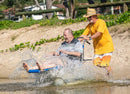 Wheeleez Sandcruiser All-Terrain Beach Wheelchair Side View In Mediterranean Blue Color With Large Balloon Wheels, Positioned In The Water With One Man Sitting In The Sandcruiser And One Man Pushing The Wheelchair Splashing Water And Smiling On A Sunny Day.