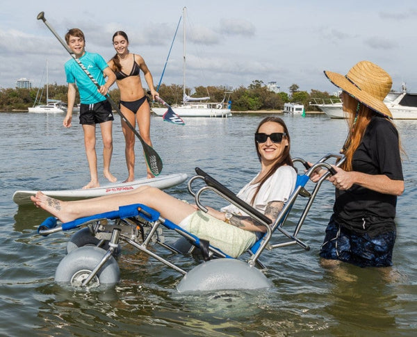 Wheeleez Sandcruiser All-Terrain Beach Wheelchair Side View In Mediterranean Blue Color With Large Balloon Wheels, Positioned In The Water With One Smiling Woman Sitting In The Sandcruiser And Woman Pushing The Wheelchair With Two Kids On A Paddle Board And Boats In The Background On A Sunny Day.