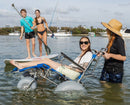 Wheeleez Sandcruiser All-Terrain Beach Wheelchair Side View In Mediterranean Blue Color With Large Balloon Wheels, Positioned In The Water With One Smiling Woman Sitting In The Sandcruiser And Woman Pushing The Wheelchair With Two Kids On A Paddle Board And Boats In The Background On A Sunny Day.