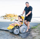 Wheeleez Sandpiper All-Terrain Beach Wheelchair Front View Yellow And Silver Color With Large Wheels Designed For Children And Small Adults On A Concrete Path Man Holding Handles Of Wheelchair With Young Girl Sitting.