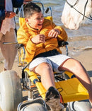 Wheeleez Sandpiper All-Terrain Beach Wheelchair Front View Yellow And Silver Color With Large Wheels Designed For Children And Small Adults On A Sandy Beach With A Young Boy Sitting And Smiling While Petting A Horse.