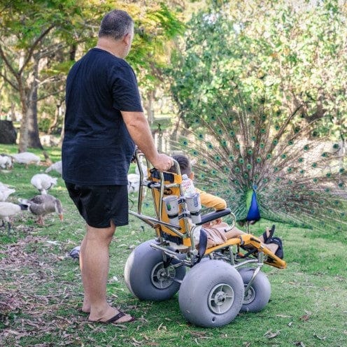 Wheeleez Sandpiper All-Terrain Beach Wheelchair Rear View Yellow And Silver Color With Large Wheels Designed For Children And Small Adults In A Green Park Setting Man Standing Behind Wheelchair With Young Boy Sitting.