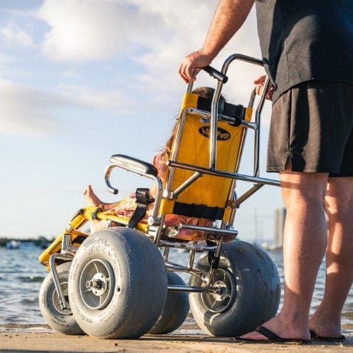 Wheeleez Sandpiper All-Terrain Beach Wheelchair Rear View Yellow And Silver Color With Large Wheels Designed For Children And Small Adults On A Sandy Beach Man Standing Behind Wheelchair With Young Girl Sitting.
